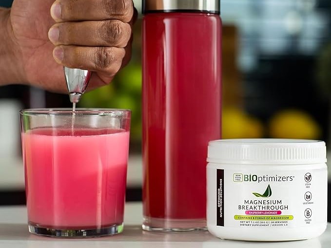 Hand frothing a pink Magnesium Breakthrough drink in a glass, with a tall bottle of the same drink and a BiOptimizers supplement tub in the foreground.